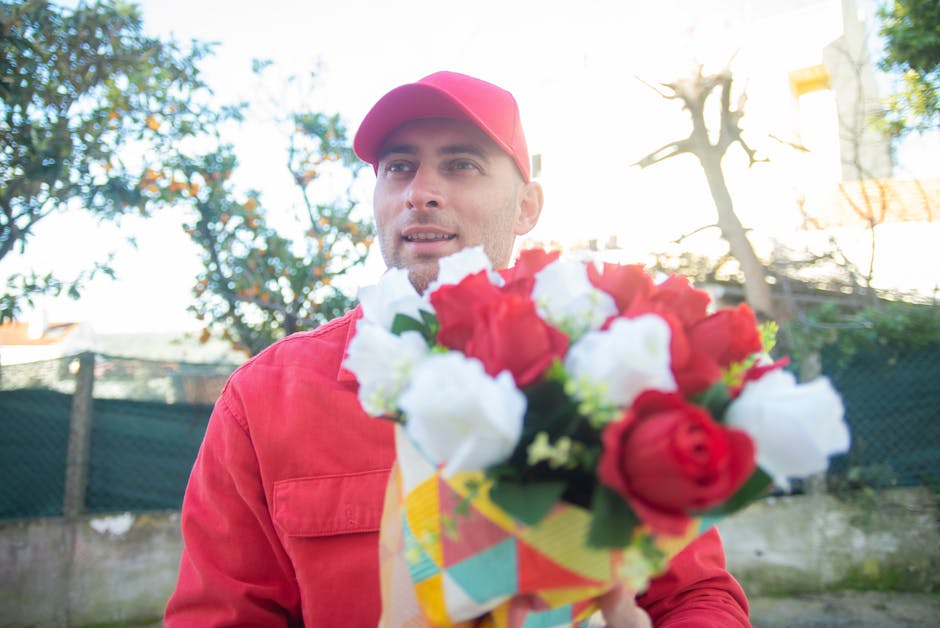 A man wearing a red cap and matching red jacket holding a vibrant floral bouquet with red and white roses, wrapped in colorful paper. He is standing outdoors in a sunny environment, with trees and a f