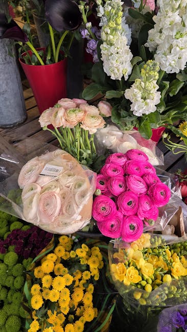 A vibrant display of fresh flowers including pink ranunculus and pale pink roses arranged in transparent wrapping, with bouquets of white stock, purple calla lilies, and yellow chrysanthemums surround