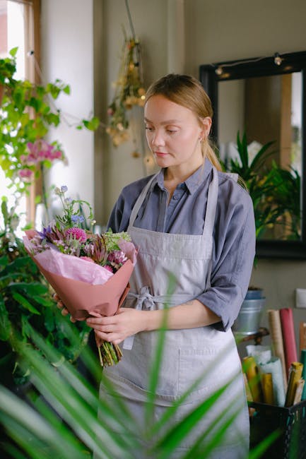 A young woman with light brown hair tied back, dressed in a blue shirt and a light grey apron, stands indoors in a floral shop, delicately holding a bouquet of fresh flowers wrapped in pink paper. The