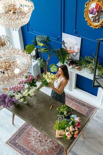 A woman with long brown hair, wearing a beige top and dark skirt, arranging a bouquet of fresh flowers on a large grey table in a bright, well-lit floral shop. The bouquet features white and green flo