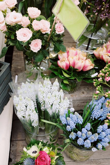 A curated display of fresh floral arrangements including pale pink roses in a glass vase, pink and yellow tulips in a clear container, and clusters of white and blue grape Hyacinths in transparent wra