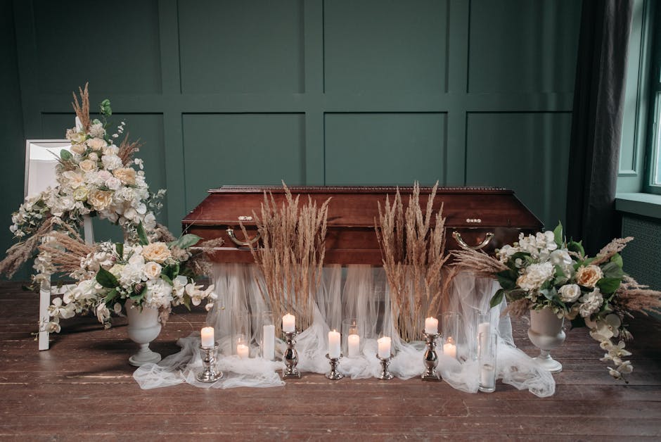 A floral tribute featuring two large white urn arrangements with roses, lilies, and greenery, flanking a vintage wooden casket situated on a polished wooden floor. The display is enhanced with tall dr