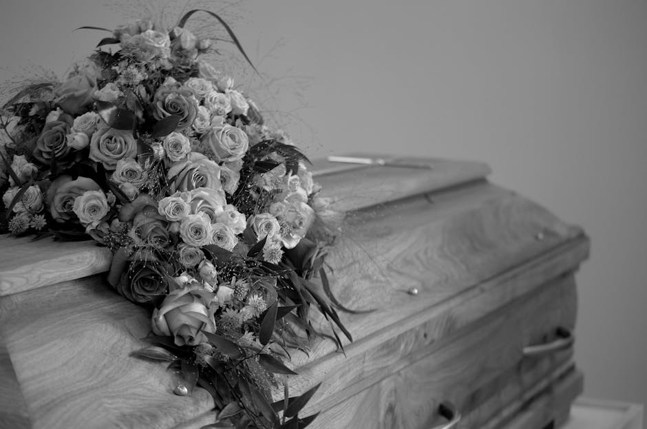 A large, somber funeral flower spray resting on a polished wooden casket. The bouquet features a variety of fresh flowers, including roses, carnations, and fillers, arranged in a cascading style. The 